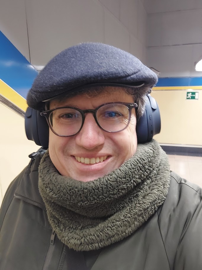 Un hombre gordo con gafas, boina negra y braga verde, con unos cascos inalámbricos azules, sonríe desde una estación de metro.