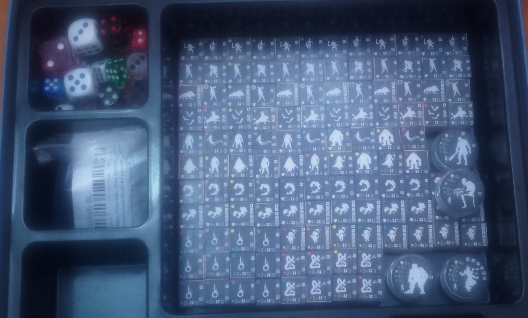 Una caja de juego estándar con insertos de plástico para organizar los componentes: en el espacio cuadrado principal pueden verse todas las fichas de monstruos, y uno de los cajetines secundarios está lleno de dados de 6 caras de diferentes tamaños y colores.