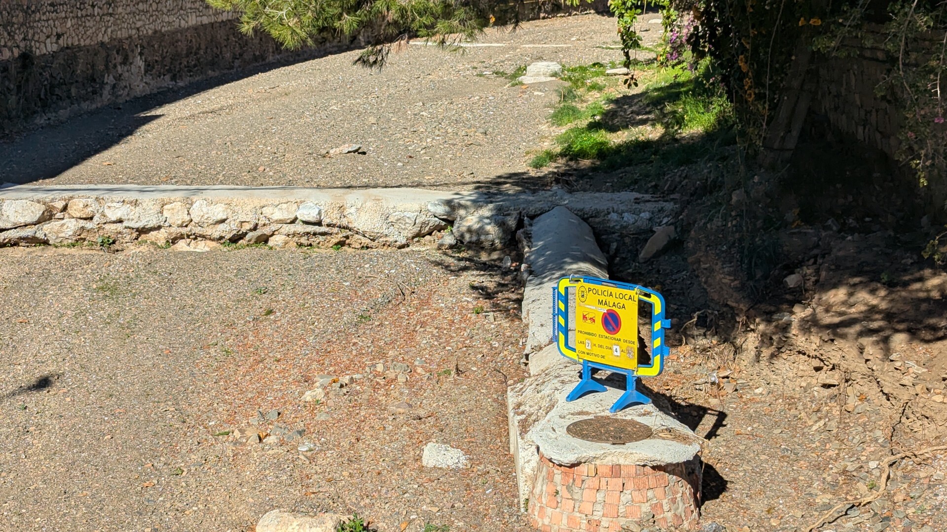 En la foto se aprecia el cauce completamente seco y terrizo de un arroyo, en el que destaca un tubo recubierto de cemento que da a una alcantarilla elevada aproximadamente medio metro sobre el suelo, todo dentro del cauce. Encima de la alcantarilla hay una valla de la Policía Local de Málaga informando de la prohibición temporal de aparcar en la zona a partir de las 7 de la mañana del día 4, sin especificar nada más.