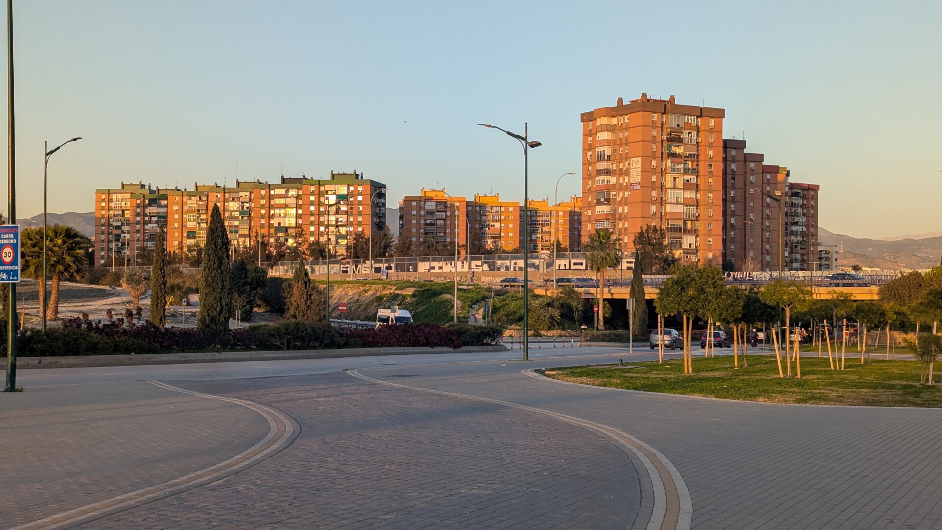 Foto urbana en la que se aprecia una calle con mediana y zonas verdes alrededor, que cruza por debajo de un puente que alberga la autovía, de la que poco se aprecia salvo algunos coches. Al fondo, varios edificios residenciales en los que está incidiendo el sol del atardecer, que está ya bastante bajo, dándoles una tonalidad más anaranjada. A la derecha de la imagen, a la lejanía, se aprecian algunas montañas.