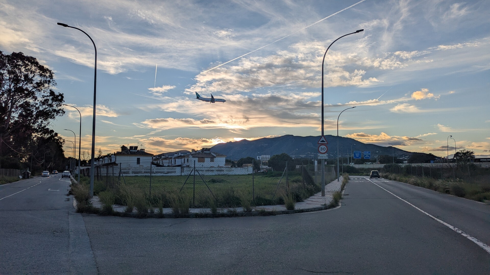 En la foto se puede apreciar un entorno residencial, poco edificado y con las pocas viviendas siendo casas mata, detrás de un pequeño prado cercado por una valla, el cual a su vez lo rodea una calle. En mitad del cielo, que está con el sol de atardecer asomando entre las nubes, destaca un avión de Transavia preparándose para aterrizar en el cercano aeropuerto de Málaga