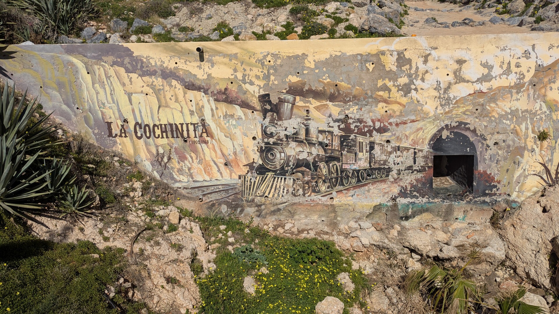 Pintado en una pared, y con bastante pintura caída del tiempo, se aprecia un dibujo de un antiguo tren de vapor saliendo de un túnel y el texto 