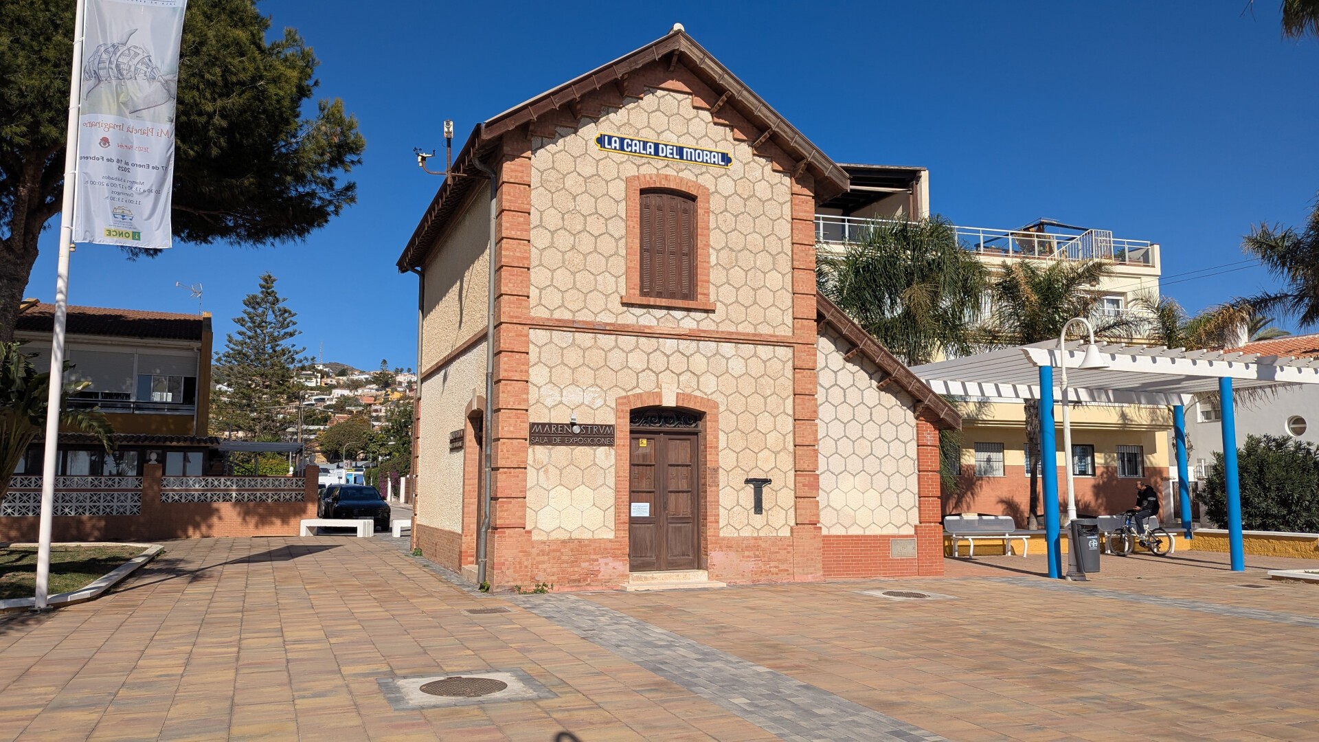 En la foto se puede apreciar una estrecha edificación de dos alturas con tejado a dos aguas en el medio de una plaza peatonal. En el frontal se aprecia una puerta de madera cerrada con un cartel 