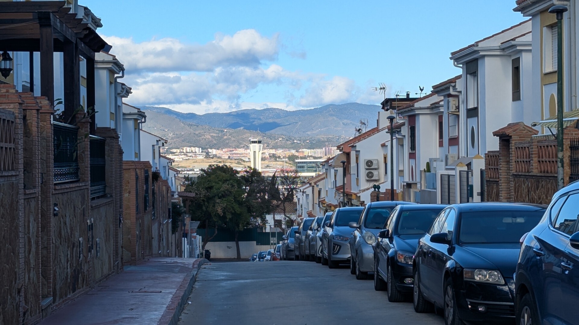 En la foto se aprecia una calle residencial, de un sólo sentido hacia el fotógrafo, con una fila de coches aparcados a la derecha y viviendas unifamiliares a ambos lados. La calle describe una pendiente descendiente, suave al inicio pero tan pronunciada que enseguida se deja de ver el resto de la calle. Al fondo, a lo lejos y detrás de un árbol, se aprecia la torre de control del Aeropuerto, indicando la cercanía del mismo de las casas. Varias edificaciones más a lo lejos y los montes de Málaga completan el paisaje, en un día con cielos azules y algunas nubes.