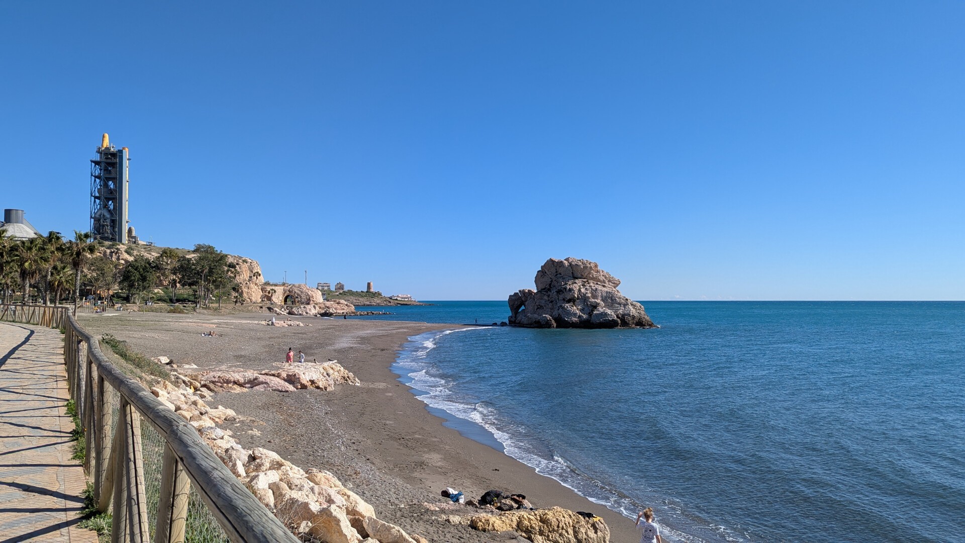 En la foto se aprecia el mar, mayormente tranquilo, a la derecha, rompiendo algunas olas contra la playa, que queda a la izquierda. Algunos bañistas, pocos, se aprecian en la playa, en la que destacan también algunas rocas. En la parte central de la foto, conectado con la playa por un istmo muy pequeño, una gran roca de unos 8 metros se adentra en el mar. Desde el punto de vista de la foto se aprecia en primera persona la barandilla de madera de un pequeño paseo, y a la izquierda de la foto, al fondo, la torre de la fábrica de cementos de la zona.